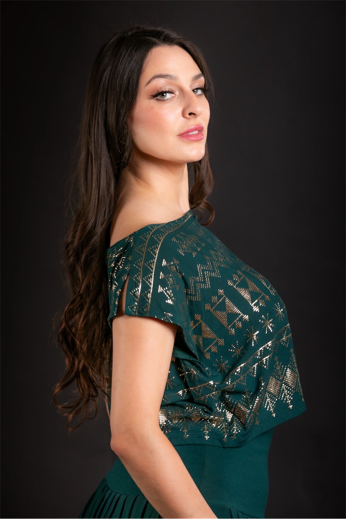 A woman with long, wavy brown hair wears the Melodia Designs Alkimiya Dede Crop Malachite with gold foil geometric patterns, standing against a black background and looking over her shoulder at the camera.