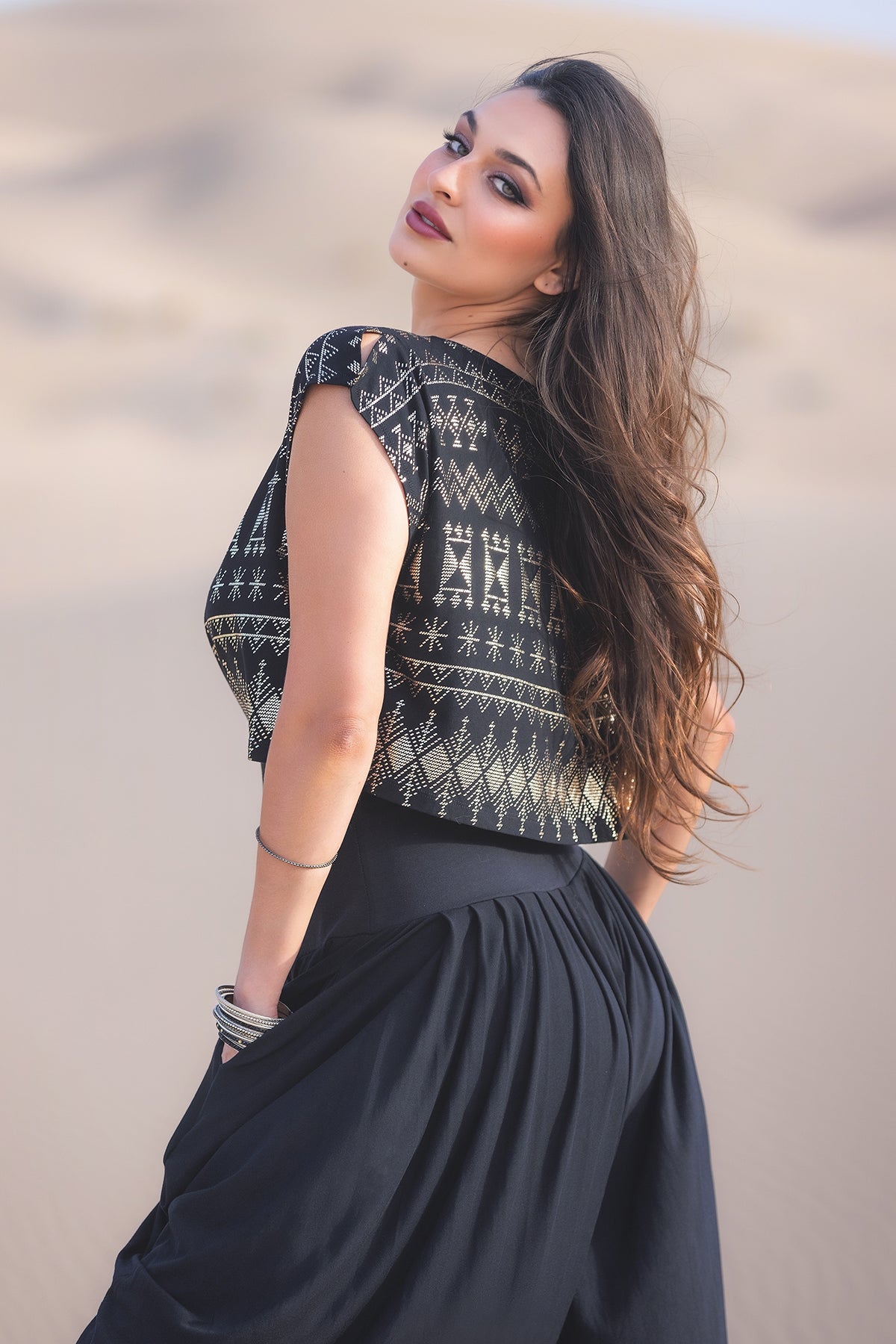 A woman with long, wavy brown hair stands in a sandy desert, wearing Melodia Designs Jazmine Harem Pant in Obsidian and a black patterned crop top, accented by bracelets on her left wrist as she looks over her shoulder.