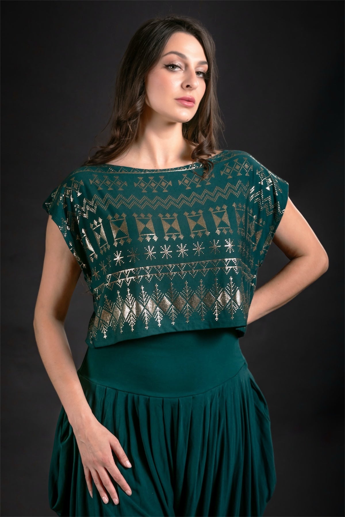 A woman poses confidently in the Alkimiya Dede Crop Malachite by Melodia Designs, a dark green, geometric-patterned top with metallic foil accents, set against a plain dark background.
