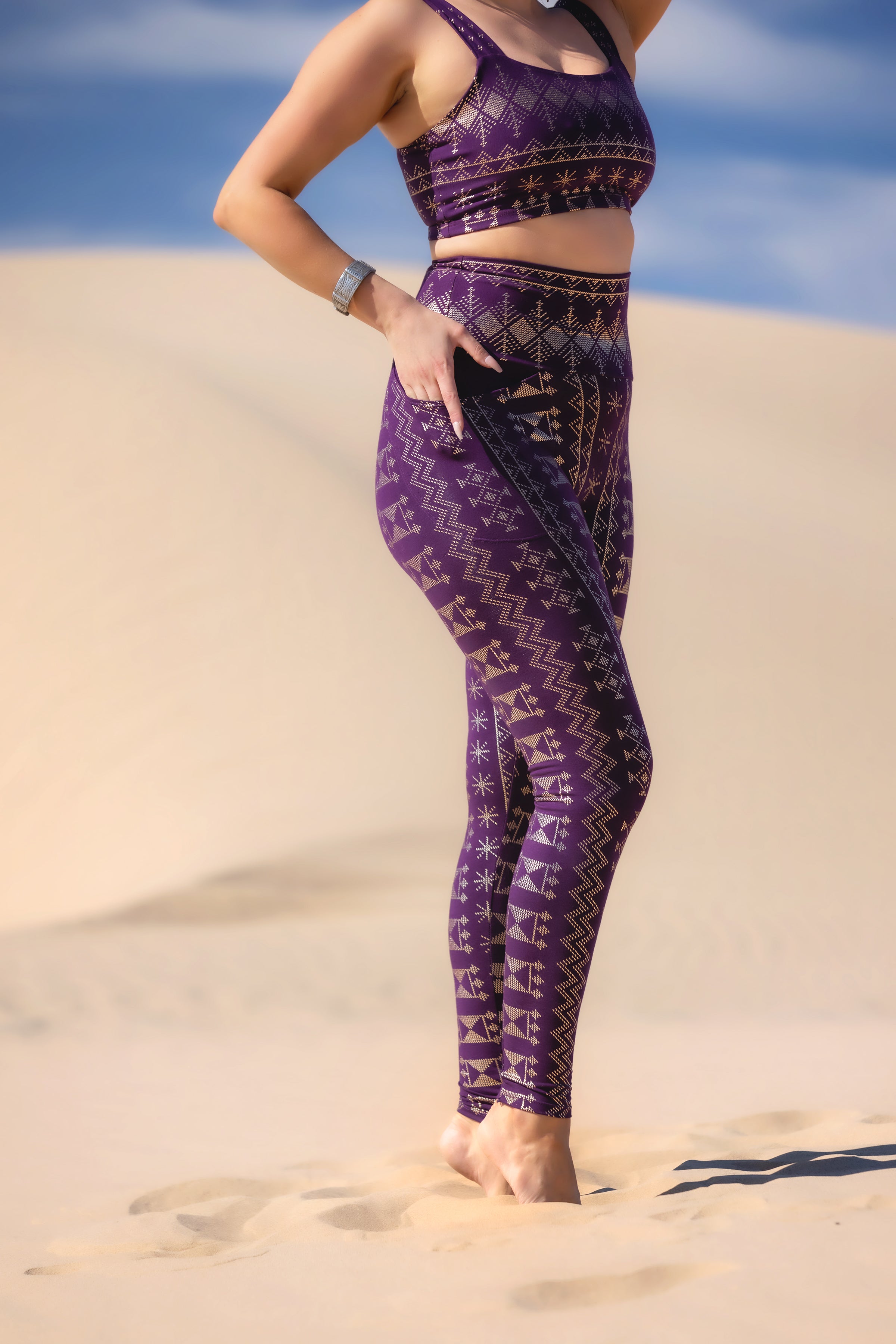 Wearing Melodia Designs' Alkimiya Elsa Legging Aubergine-Gold v2, a person stands barefoot on sand dunes with one hand on their hip; face is out of frame.