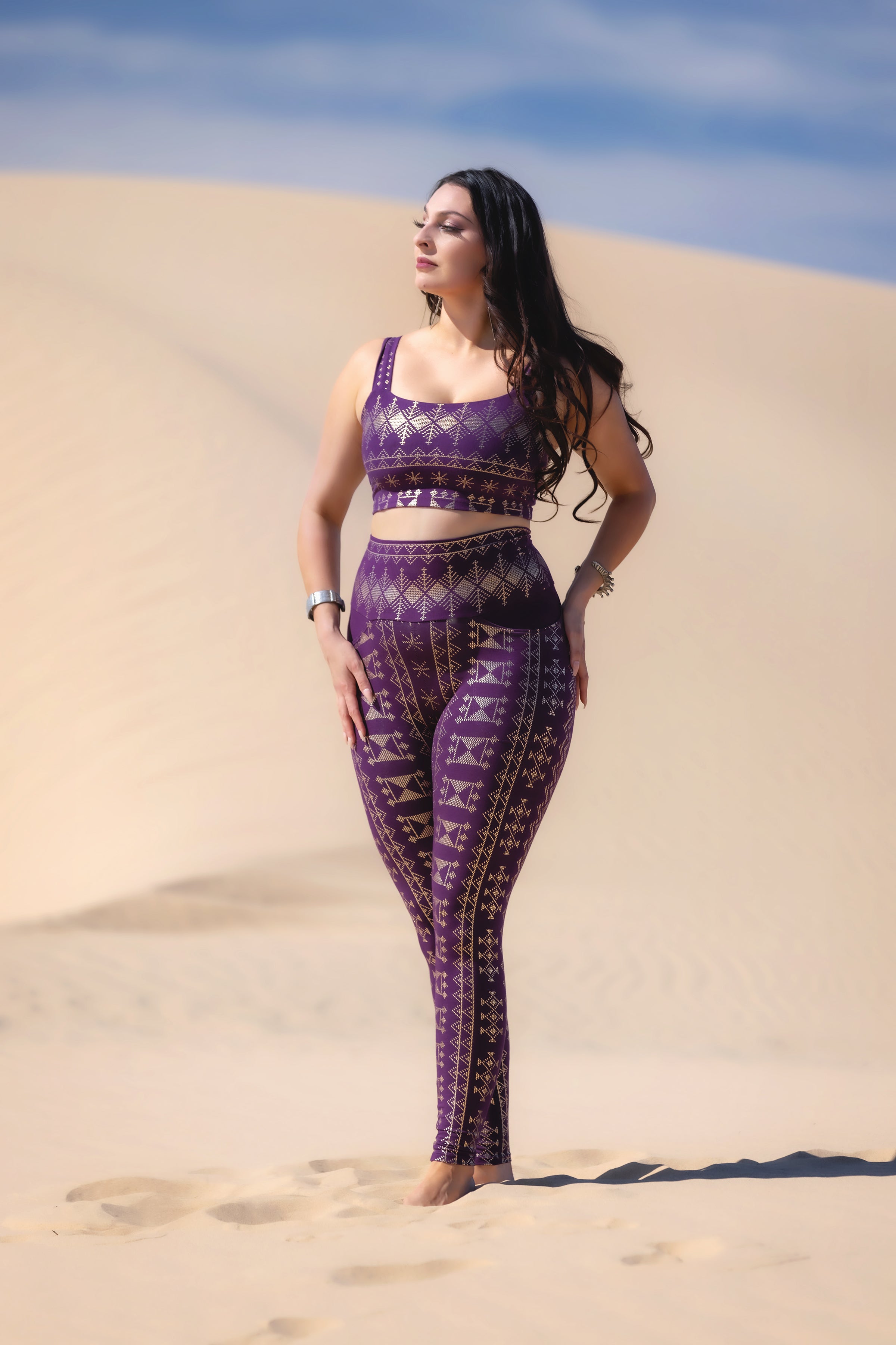 A woman with long dark hair stands on sand dunes, wearing Melodia Designs' Alkimiya Elsa Legging Aubergine-Gold v2. She looks to the side with a relaxed expression under a blue sky.