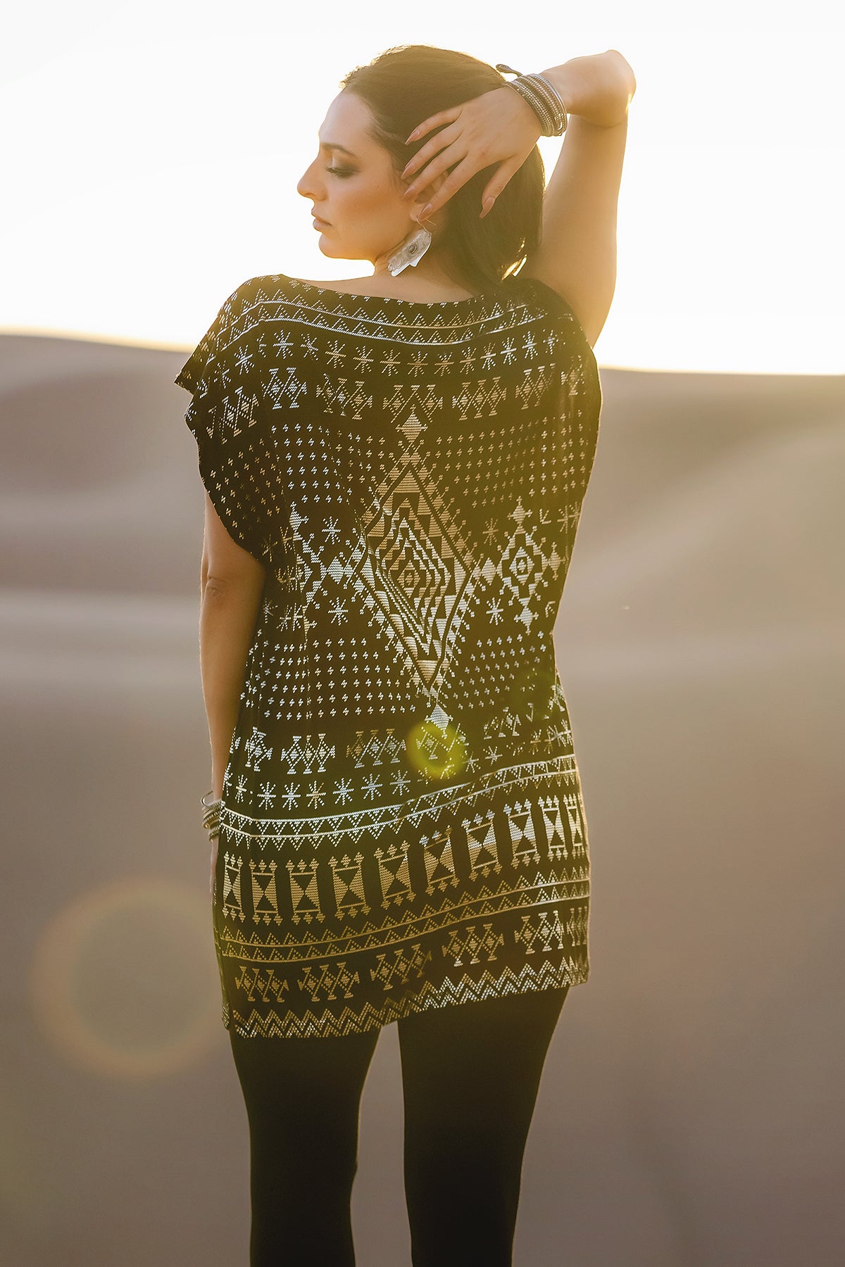 A woman stands outdoors with her back to the camera, wearing the Melodia Designs Alkimiya Jade Tunic Obsidian and black leggings. She raises one arm behind her head as sunlight highlights the tunic’s gold geometric patterns and flattering silhouette.