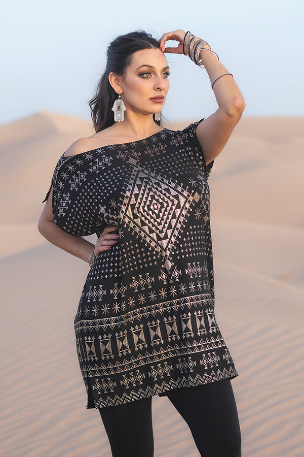 A woman stands in a sandy desert wearing the Alkimiya Jade Tunic Obsidian by Melodia Designs—an eco-friendly black tunic with geometric patterns—paired with black leggings, silver earrings and bracelets, her hair tied back as she gazes into the distance.
