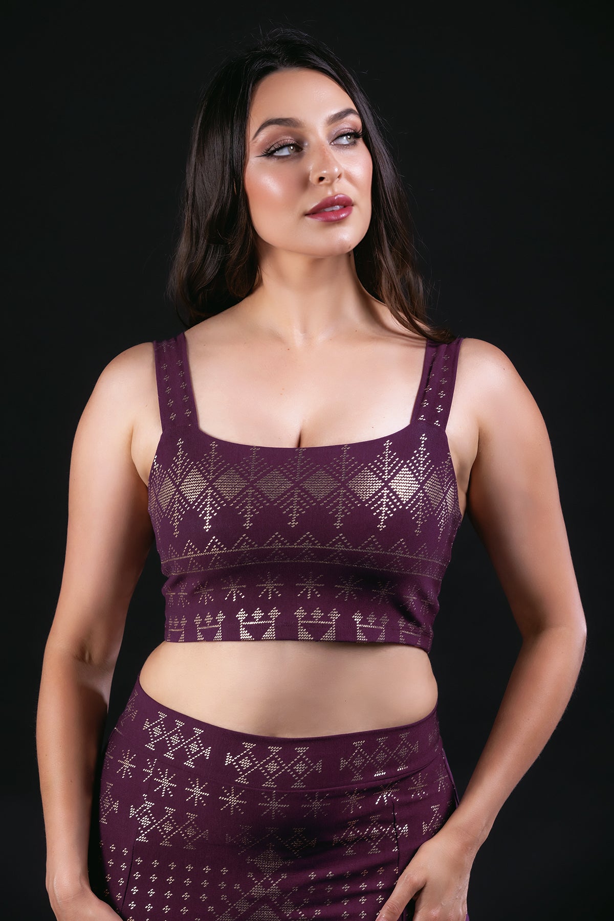 Wearing the Melodia Designs Alkimiya Neisha Bra in Aubergine-Gold, a woman stands confidently against a black background, looking slightly to the side.