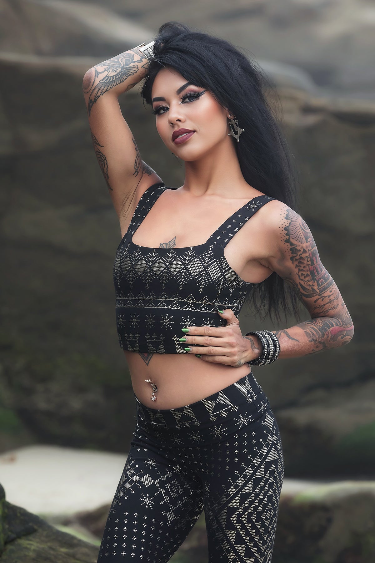 A woman with long black hair and tattoos poses outdoors on large rocks, wearing the Melodia Designs Alkimiya Neisha Bra Obsidian under her matching black and silver set, her raised arm highlighting jewelry and piercings.