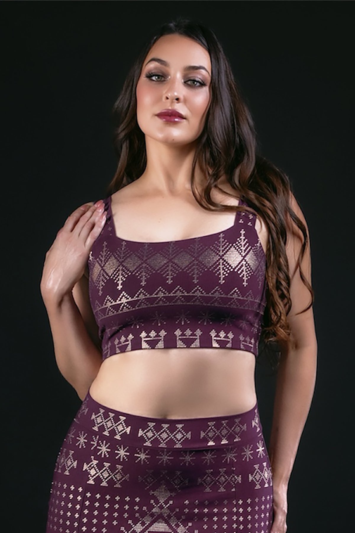 A woman with long brown hair wears the Alkimiya Neisha Bra Aubergine-Gold by Melodia Designs, featuring eco-luxe fabric and metallic geometric patterns, posing against a black background.