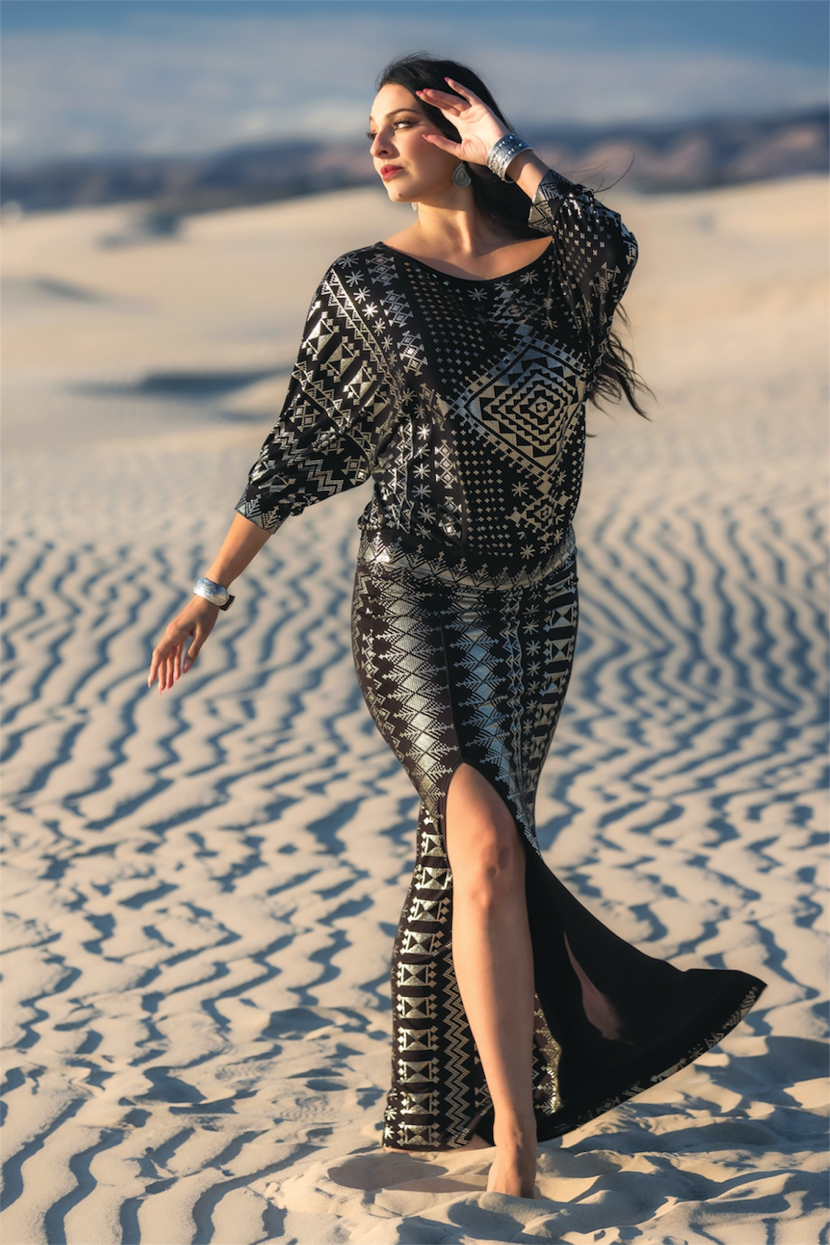 A woman in a Melodia Designs Alkimiya Vanessa Skirt Obsidian-Antique Silver, barefoot on sunlit sand dunes with her hair blowing and arm raised, is a stunning vision of sustainable fashion.