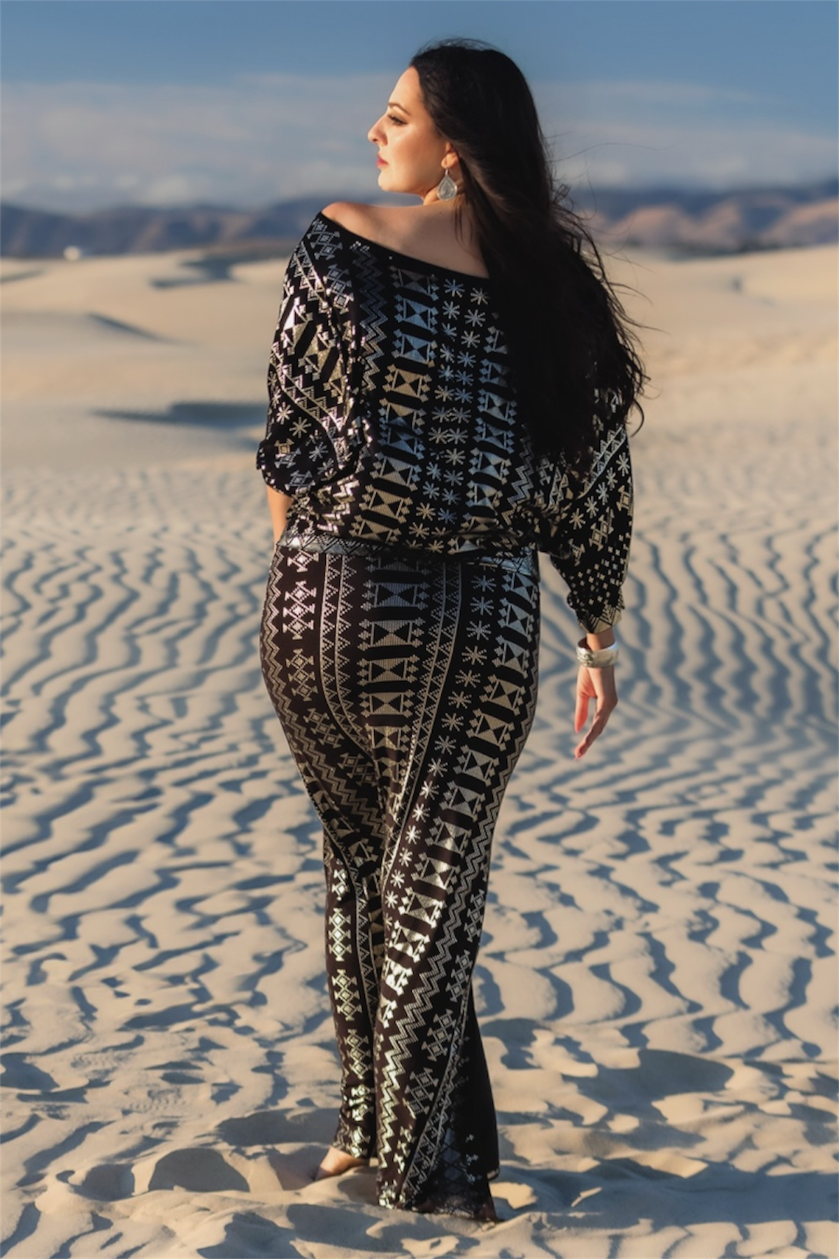 A woman with long dark hair walks barefoot on sandy dunes beneath a clear sky, wearing the Alkimiya Vanessa Skirt Obsidian-Antique Silver by Melodia Designs—embracing sustainable fashion with distant mountains behind her.