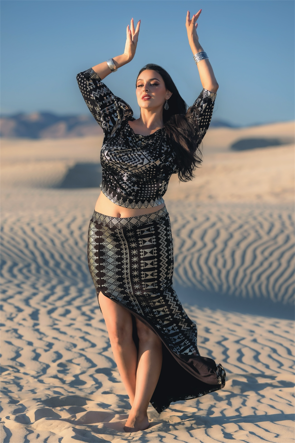 A woman poses with raised arms in a sandy desert, wearing the Melodia Designs Alkimiya Bella Batwing Blouse in Obsidian-Antique Silver, featuring an eco-chic patterned black and silver off-the-shoulder design.