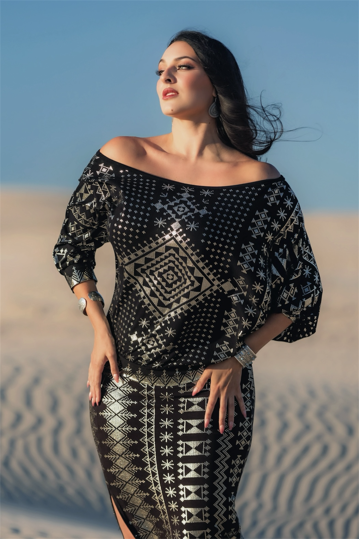 A woman in a Melodia Designs Alkimiya Bella Batwing Blouse in Obsidian-Antique Silver stands in a sandy desert, gazing sideways with one hand on her hip. She wears her dark hair loose, accessorized with earrings and a bracelet under a clear blue sky.