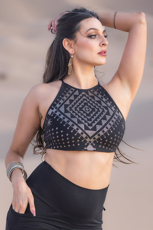 A woman with long, dark hair poses confidently outdoors in the Melodia Designs Alkimiya Olga Pyramid Bra Obsidian crop top and matching skirt, paired with earrings and bangles against a blurred sandy background.