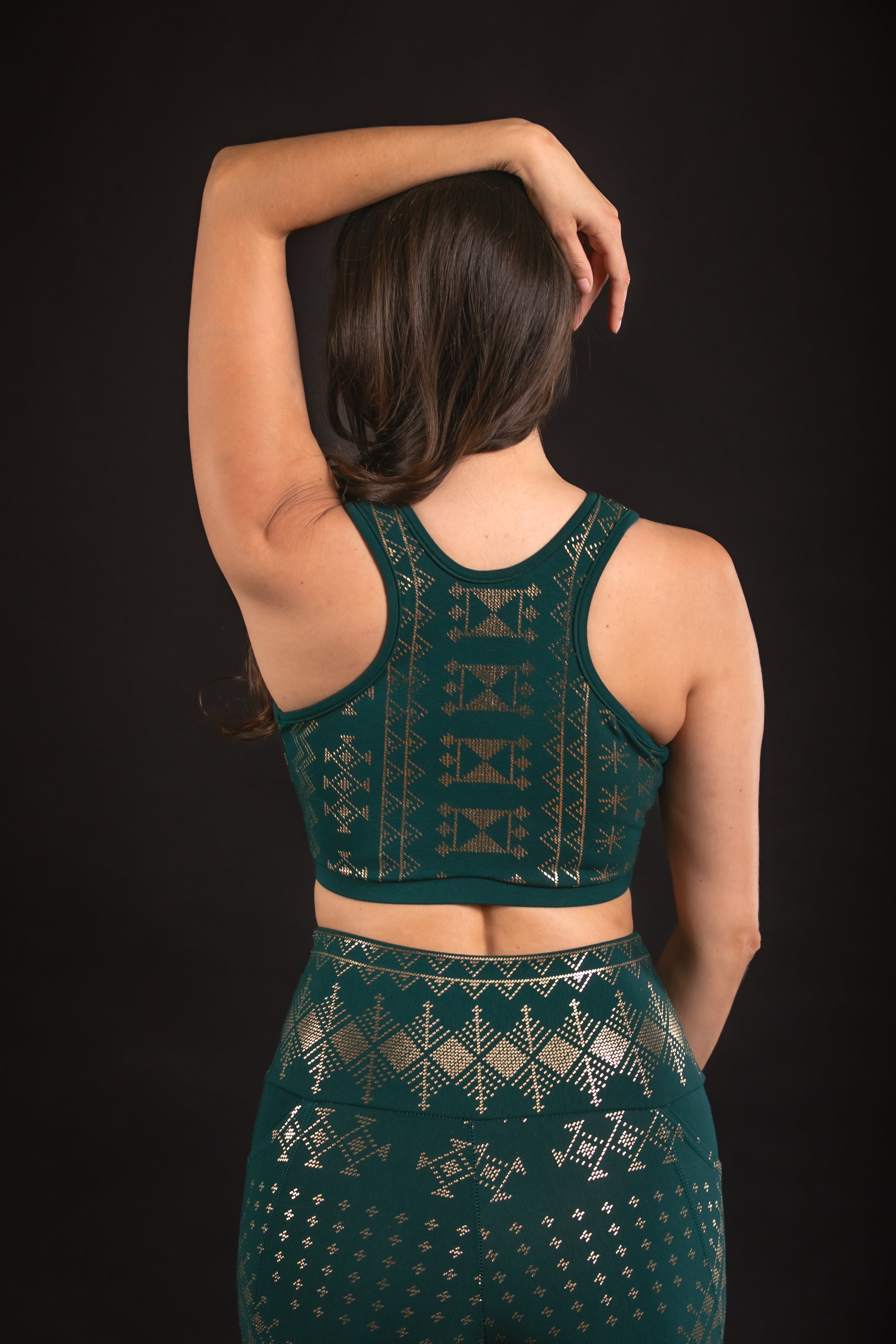 A woman with long brown hair poses with one arm raised, showcasing the eco-friendly Alkimiya Rainah Bra Malachite by Melodia Designs, featuring metallic geometric patterns, against a black background.