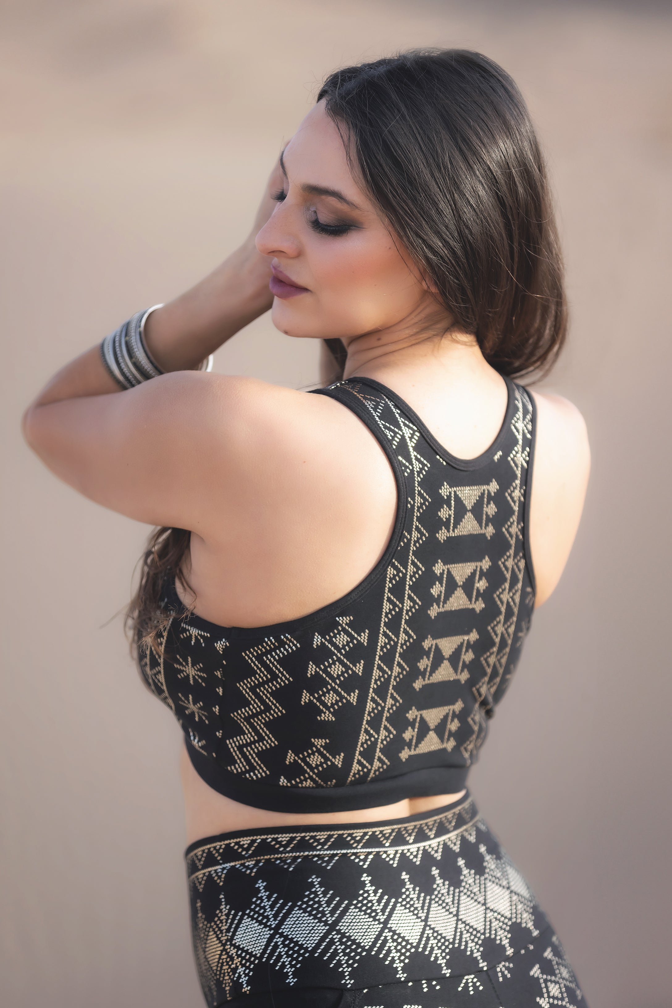 A woman with long dark hair wears the eco-friendly Melodia Designs Alkimiya Rainah Bra Obsidian, featuring gold geometric patterns, posing outdoors with her eyes closed and one hand in her hair.