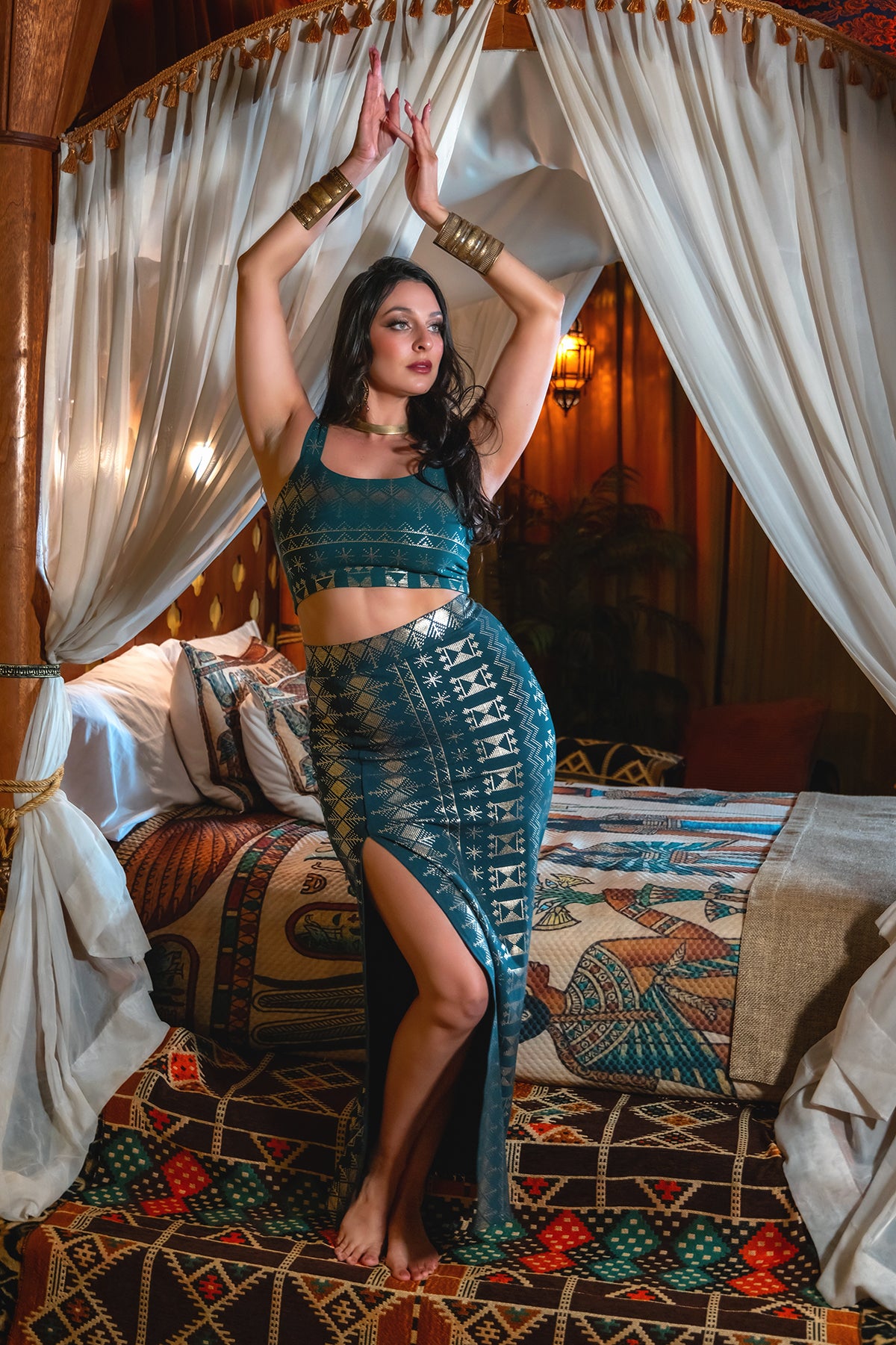 A woman wearing the Melodia Designs Alkimiya Vanessa Skirt Malachite-Gold poses with raised arms in a luxurious bedroom adorned with white drapes, rich textiles, and warm lighting.