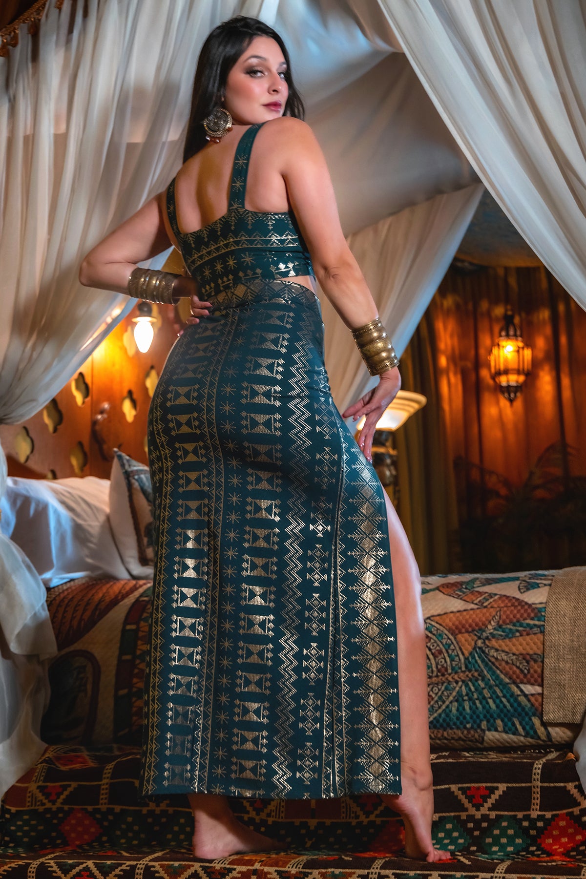 A woman stands barefoot on a bed, posing confidently in the Melodia Designs Alkimiya Vanessa Skirt Malachite-Gold. She pairs it with bold earrings and bangles, set against ornate drapes and warm, decorative lighting.