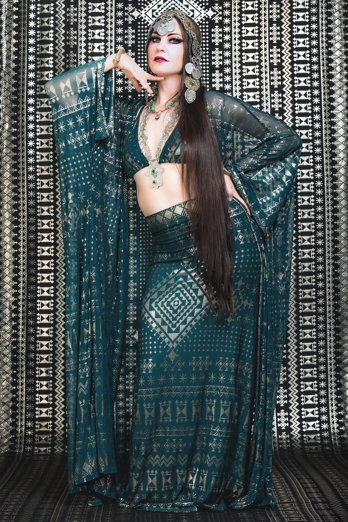 A woman models the Melodia Designs Alkimiya Zsa Zsa Robe Malachite—a teal and gold statement piece with flared sleeves and a matching headpiece—against a black-and-white geometric background, her long dark hair over one shoulder.