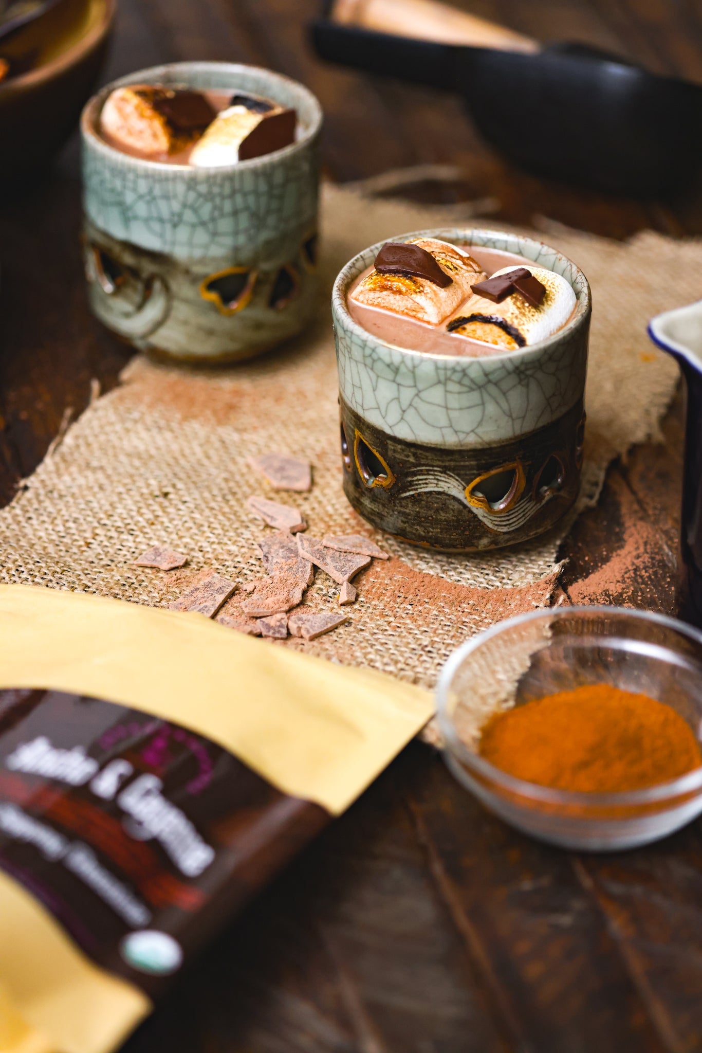 Two rustic ceramic mugs filled with chocolita.com’s Ancho & Cayenne Sipping Chocolate, topped with toasted marshmallows and chocolate pieces, rest on burlap beside cinnamon powder, chocolate shavings, and a cocoa mix packet.