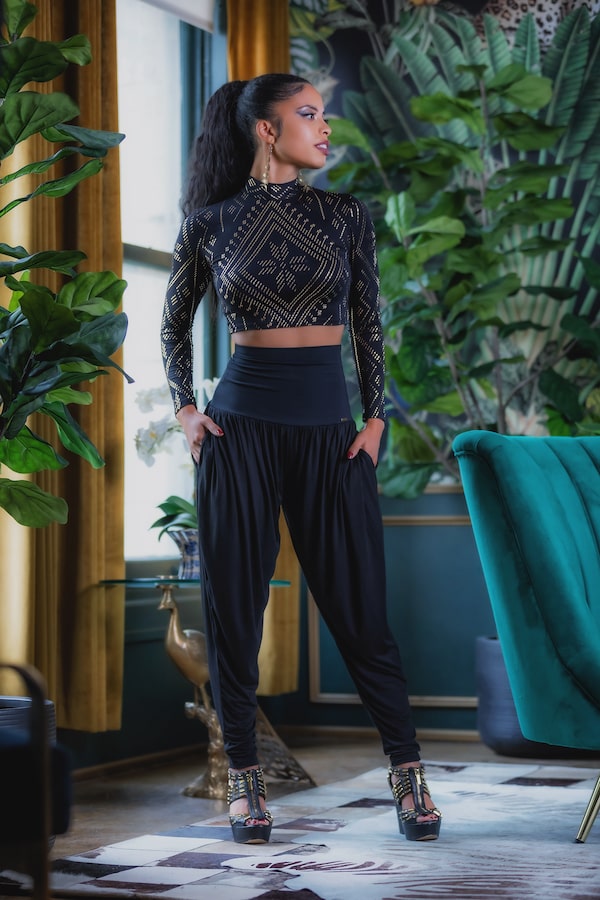 A woman wearing the Melodia Designs Auberon Mock Crop Luxor stands confidently indoors among lush plants, gold curtains, and teal furniture.