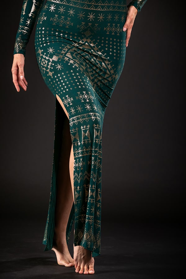 A person stands barefoot against a dark background, wearing the lower half of the Melodia Designs Alkimiya Nawal Dress Malachite—a long green dress with gold geometric patterns and a thigh-high slit. Only their lower body is visible.