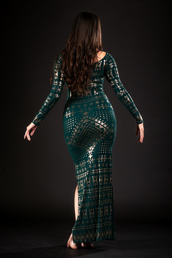 A woman with long brown hair stands barefoot, facing away, wearing the Alkimiya Nawal Dress Malachite by Melodia Designs—a long-sleeved, form-fitting green dress with gold geometric patterns against a dark background.