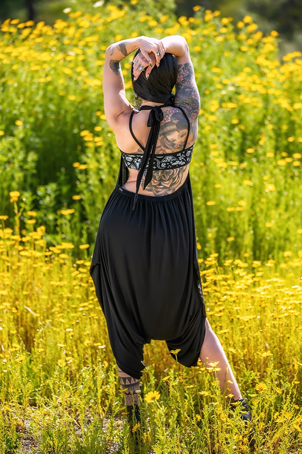 A tattooed person wearing the Melodia Designs Briana Jumper 2.0 in eco-luxe modal stands in a field of yellow wildflowers, facing away with arms raised and hands clasped, enjoying the sunny outdoors.