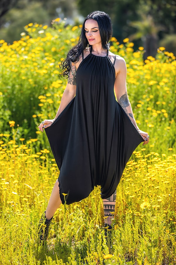 A woman with long dark hair and tattoos wears a loose black Melodia Designs Briana Jumper 2.0 while standing in a sunlit field of yellow wildflowers, softly smiling as she holds out the sides of her outfit.