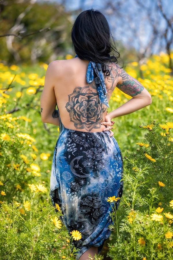 A tattooed woman stands in a field of yellow flowers, facing away and surrounded by greenery and sunshine. She wears the Melodia Designs Briana Jumper Tie Dye Celestial, featuring a celestial-inspired print.