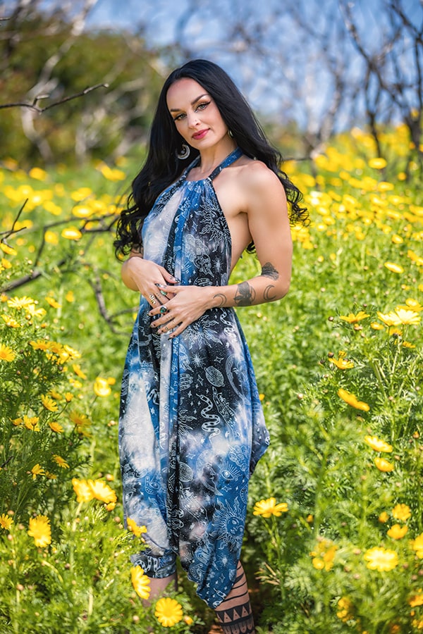 A woman with long black hair stands in a sunny field wearing the Melodia Designs Briana Jumper Tie Dye Celestial; hoop earrings sparkle, tattoos show on her arm, and she gazes at the camera with a slight smile.