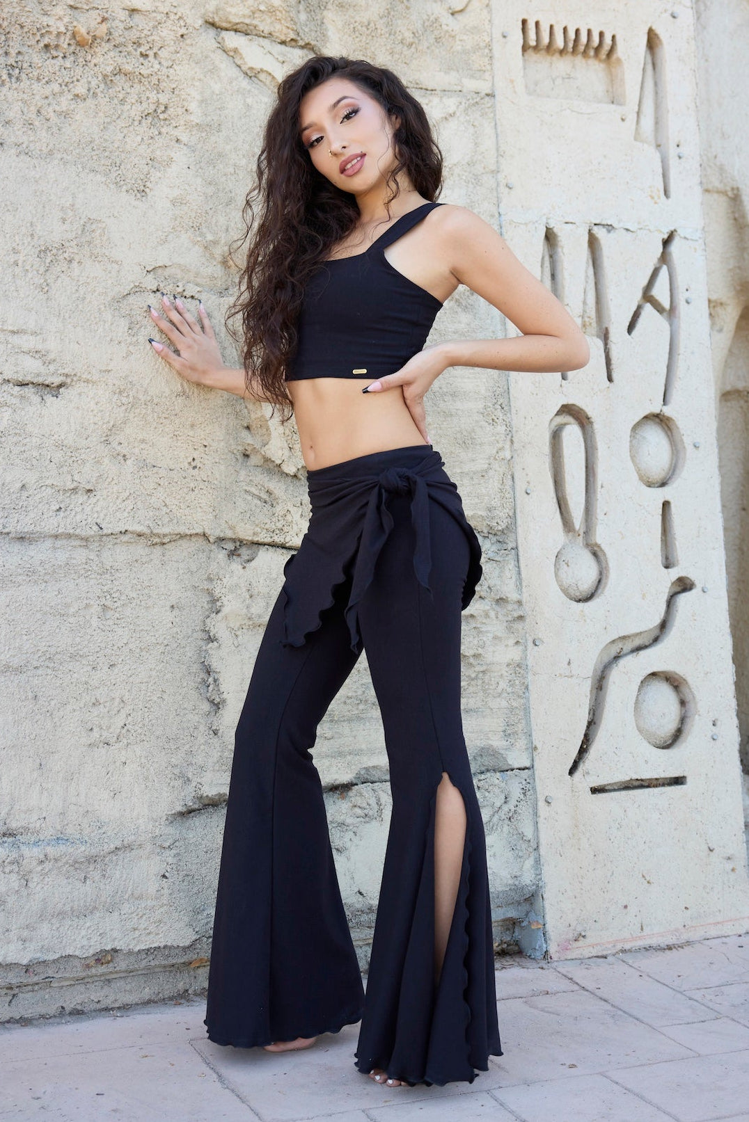 A woman posing in a black two-piece activewear set consisting of a halter crop top and high-waisted flared pants with side slits, standing against a textured stone wall with ancient hieroglyphic carvings.