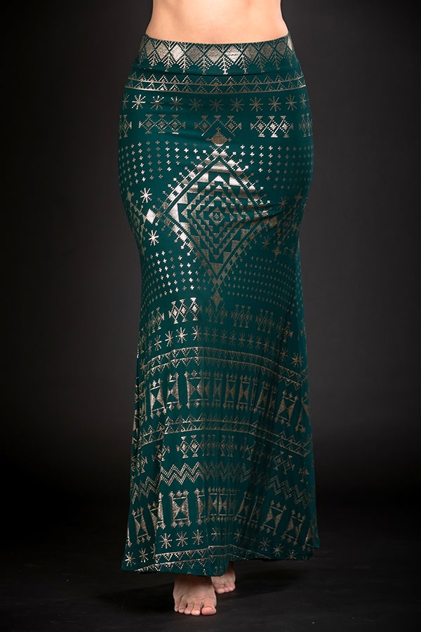 A person stands barefoot wearing the Melodia Designs Alkimiya Cleopatra Maxi Skirt in Malachite, featuring eco-luxe fabric with intricate gold geometric and tribal patterns on a dark green and black background. Only the lower torso and legs are visible.