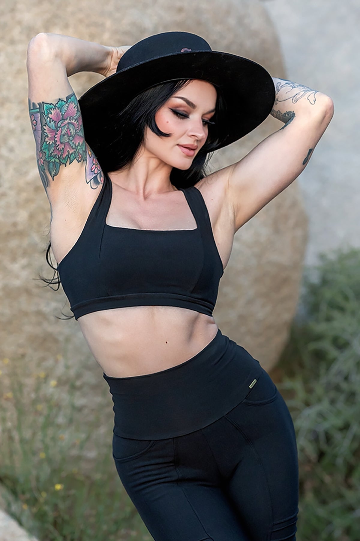 A woman with long dark hair and floral arm tattoos poses outdoors among desert rocks and plants, wearing Melodia Designs Serena Bra 2.0 in black with high-waisted leggings made from eco-luxe fabric, her arms raised confidently.