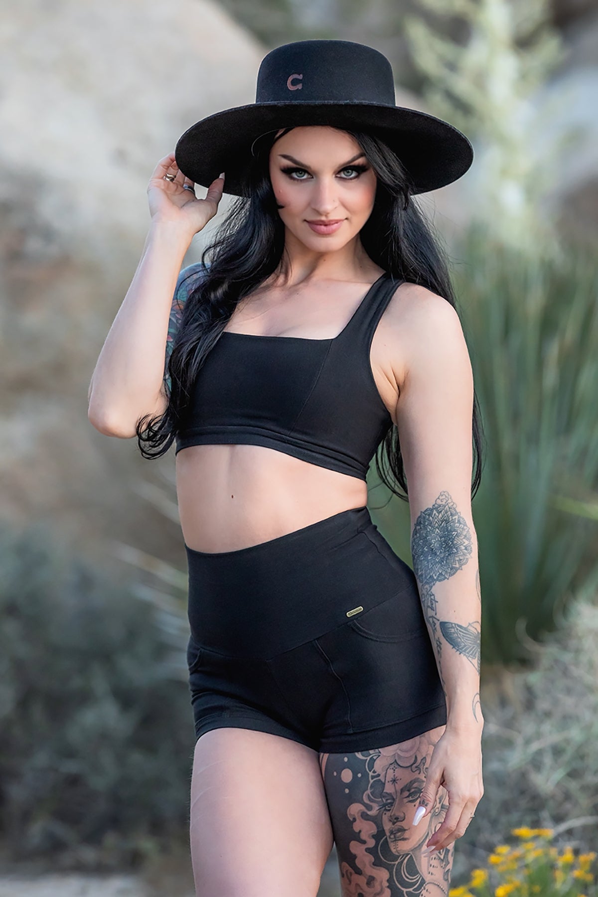 A woman with long black hair poses outdoors in the Serena Bra 2.0 by Melodia Designs—crafted from eco-luxe fabric—styled with high-waisted shorts and a wide-brimmed hat, highlighting her arm and leg tattoos among rocks and greenery.