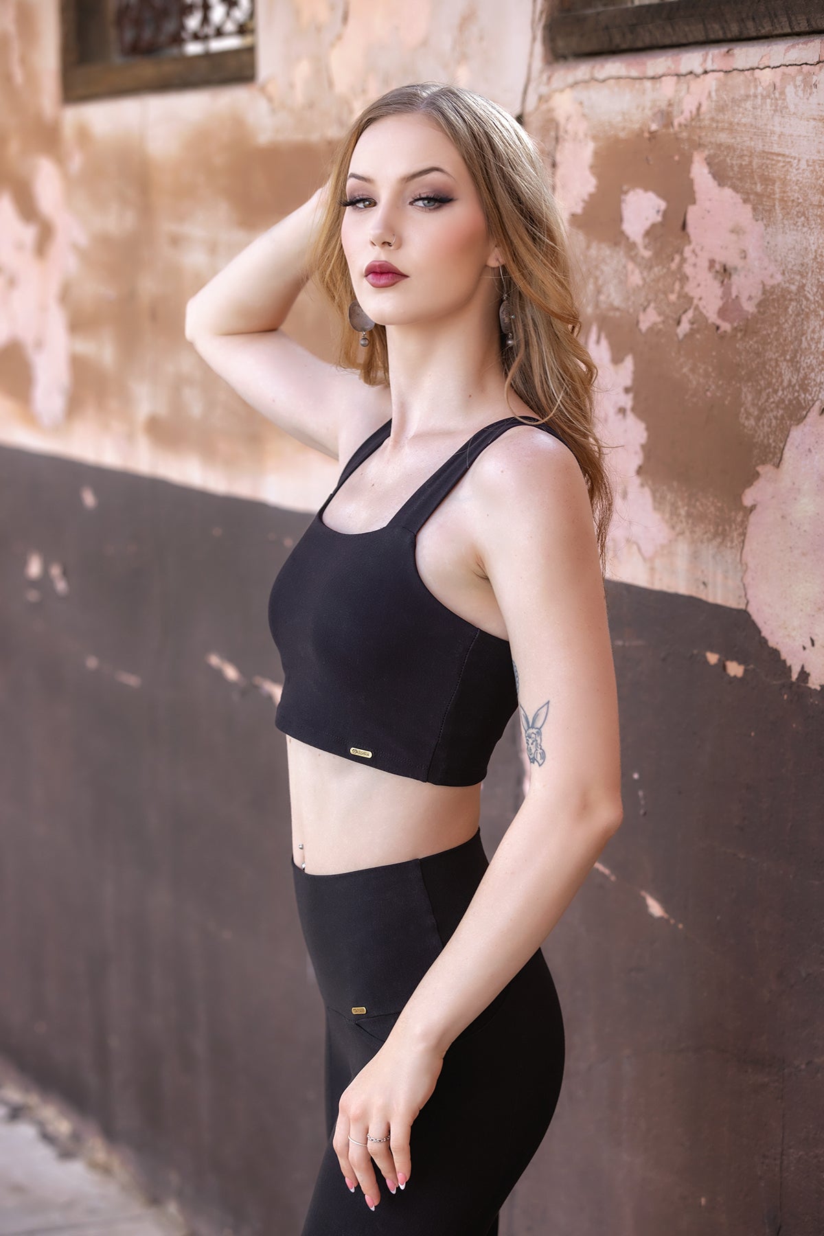 A woman in a black supportive bra and leggings stands against a weathered, pink and brown wall, posing with one hand in her hair. She has long blonde hair and a small tattoo on her upper left arm. The eco-luxe fabric of her Melodia Designs Neisha Bra adds both comfort and style to her effortlessly chic look.