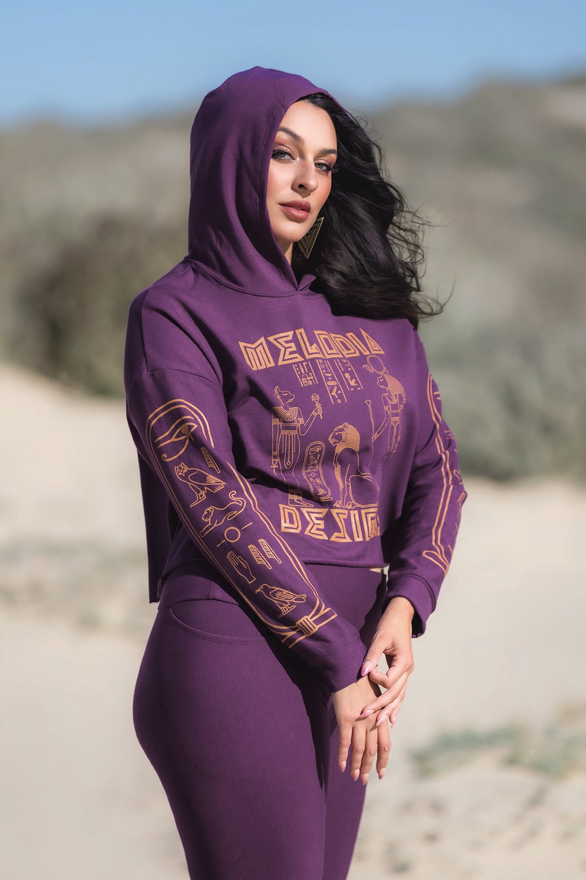 A woman with long dark hair stands outdoors on sand, wearing the Melodia Designs Crop Hoodie Fleece Aubergine and matching pants, featuring ancient Egyptian graphics and “MELODIA DESIGN” text. She poses with her hands clasped.