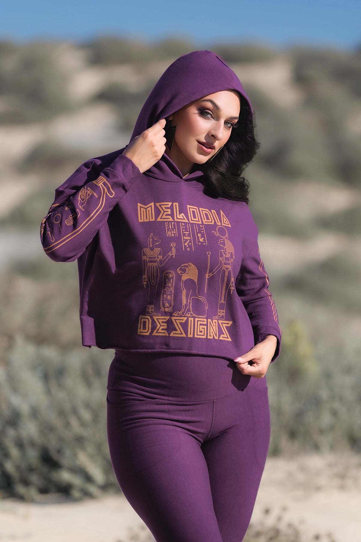 A woman stands outdoors in a Melodia Designs Crop Hoodie Fleece Aubergine with gold art, paired with matching leggings. The greenery and hills behind her are softly blurred.