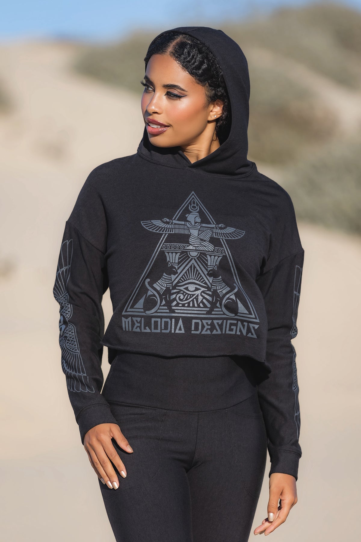 A woman stands outdoors in a black Crop Hoodie Fleece by Melodia Designs, featuring an intricate geometric design and the brand name on the front, with sandy natural scenery blurred in the background.