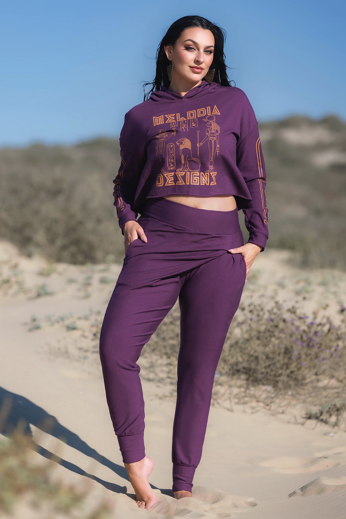 A woman stands barefoot on sand, wearing the Melodia Designs Crop Hoodie Fleece in Aubergine with a graphic design. Shrubbery and blurred dunes appear in the daylight background.