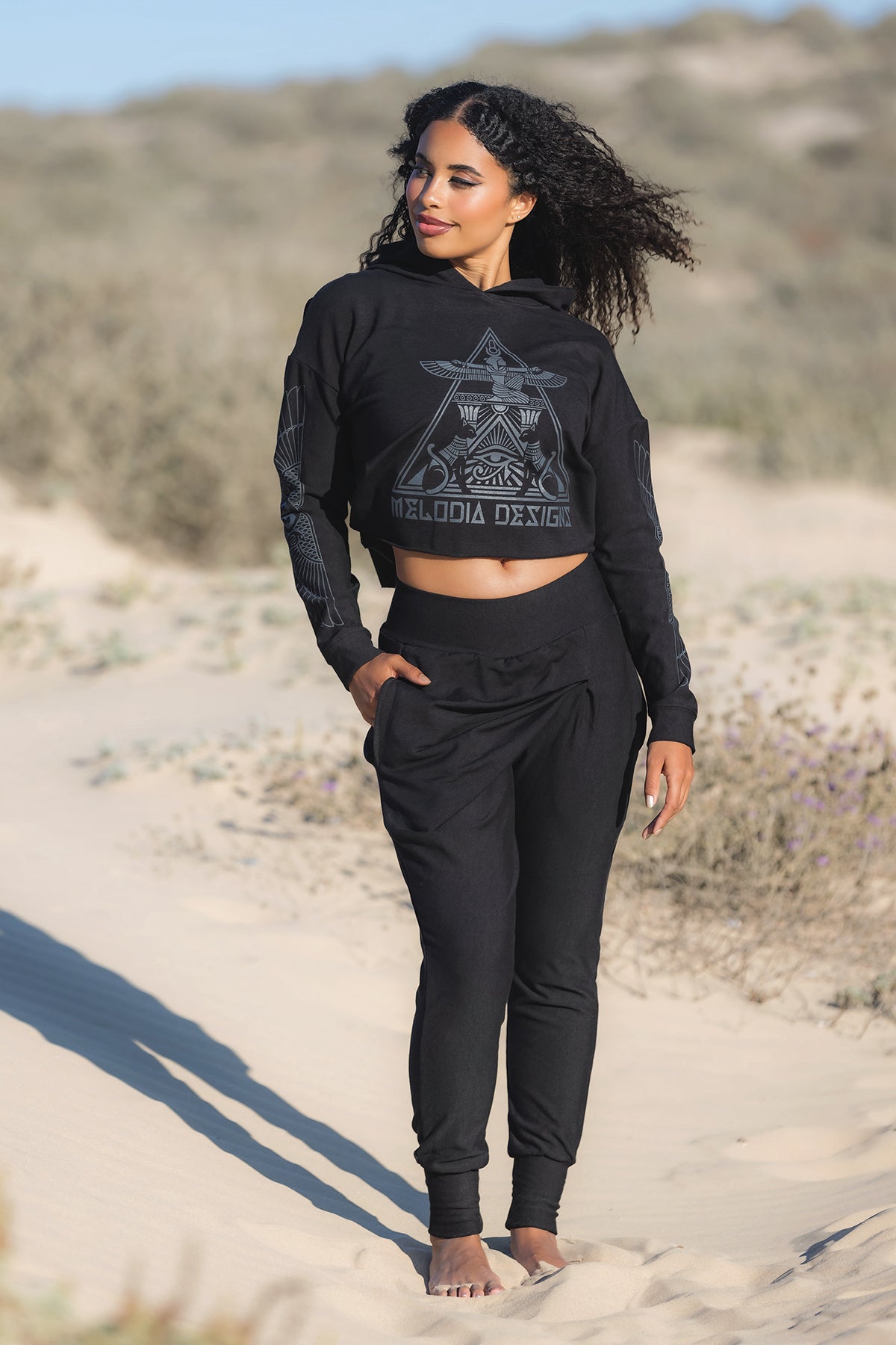 A woman stands barefoot on sandy terrain, wearing the Melodia Designs Crop Hoodie Fleece Black. Her dark curly hair blows in the wind as she gazes to the side, with dunes and sparse vegetation in the background.