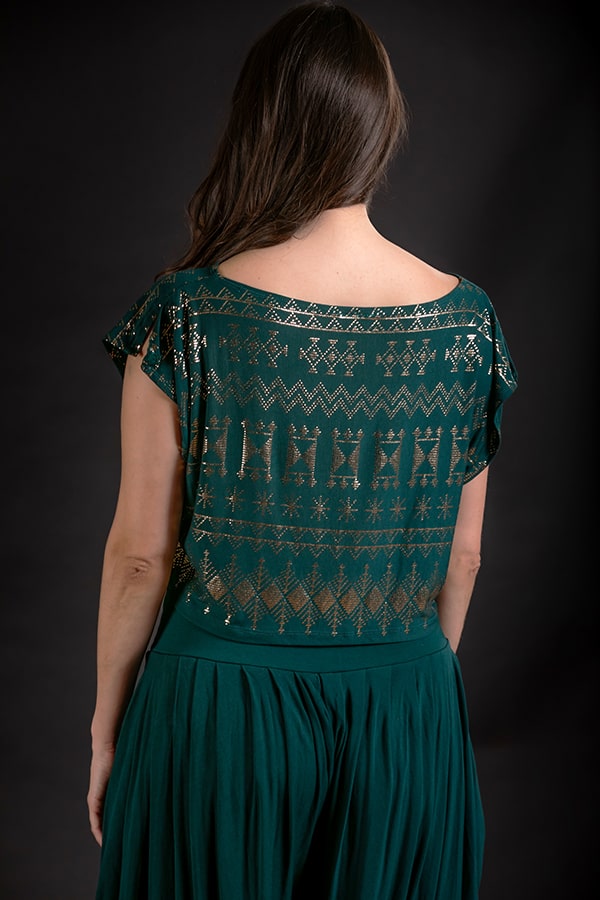 A woman with long brown hair faces away, wearing the Alkimiya Dede Crop Malachite in eco-luxe fabric with gold foil print and a matching green skirt by Melodia Designs, set against a dark background.