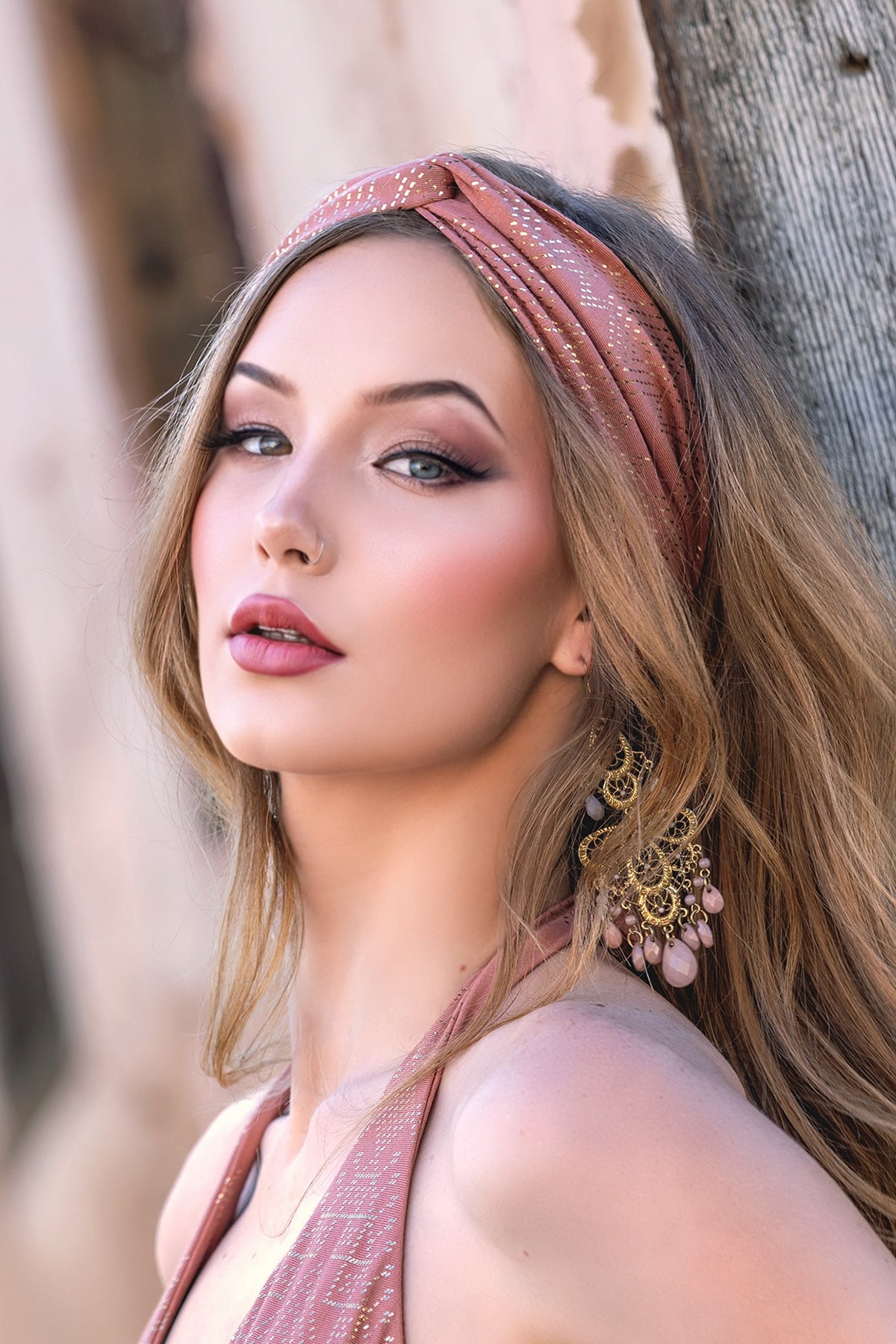 A woman with long, wavy blonde hair wears the Melodia Designs Dendera Beauty Band Desert Rose, striking bold makeup and ornate gold earrings with pink accents as she gazes confidently outdoors by a textured wall.