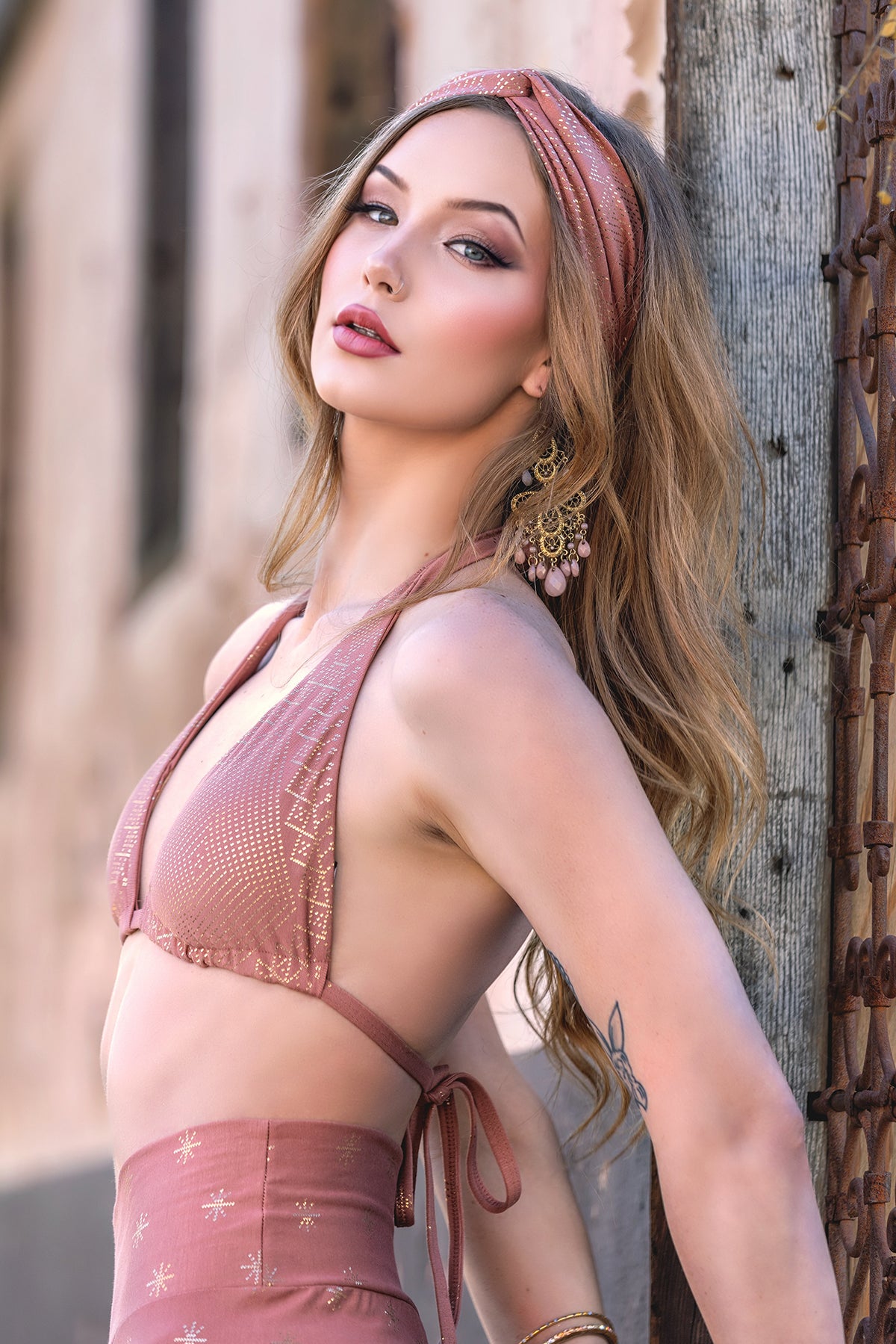 A woman with long wavy hair leans against a wooden post outdoors, wearing the Dendera Hannah Halter Desert Rose by Melodia Designs with matching bottoms. She accessorizes with a headband and large earrings, gazing confidently at the camera.