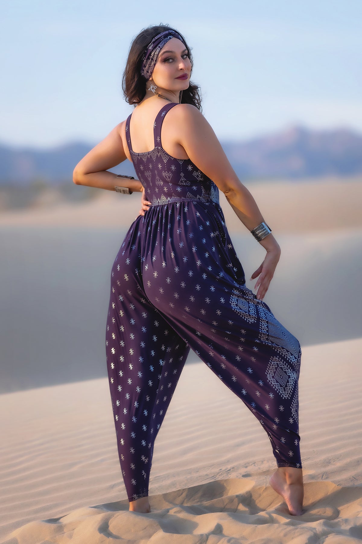 In the desert, a woman wears the Melodia Designs Dendera Jinni Jumper 2.0 Midnight Silver, featuring navy blue fabric with white star patterns and relaxed harem pant styling. She accessorizes with silver jewelry and a matching headband, gazing over her shoulder.