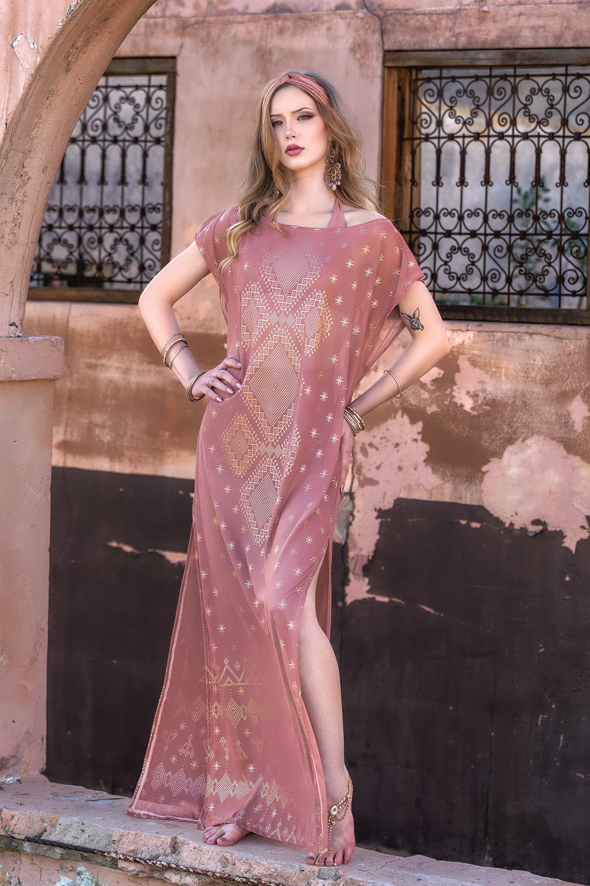 A woman stands barefoot under an archway, wearing the Melodia Designs Dendera Nadia Maxi Dress in Desert Rose, featuring gold star details. She poses against a weathered peach wall, embodying effortless resort wear elegance.