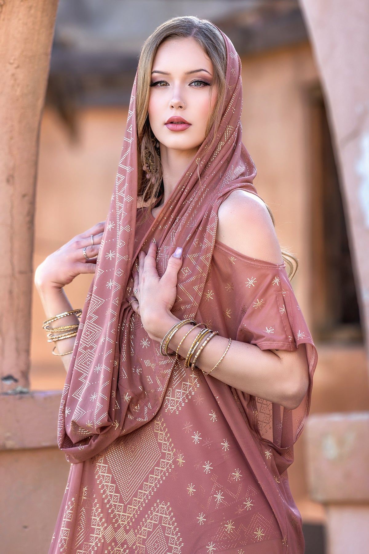A woman with light skin and long hair wears the Melodia Designs Dendera Infinity Scarf in Desert Rose over her head and shoulders. She poses outdoors in a pink patterned outfit, accessorized with gold bangles and rings, gazing at the camera.