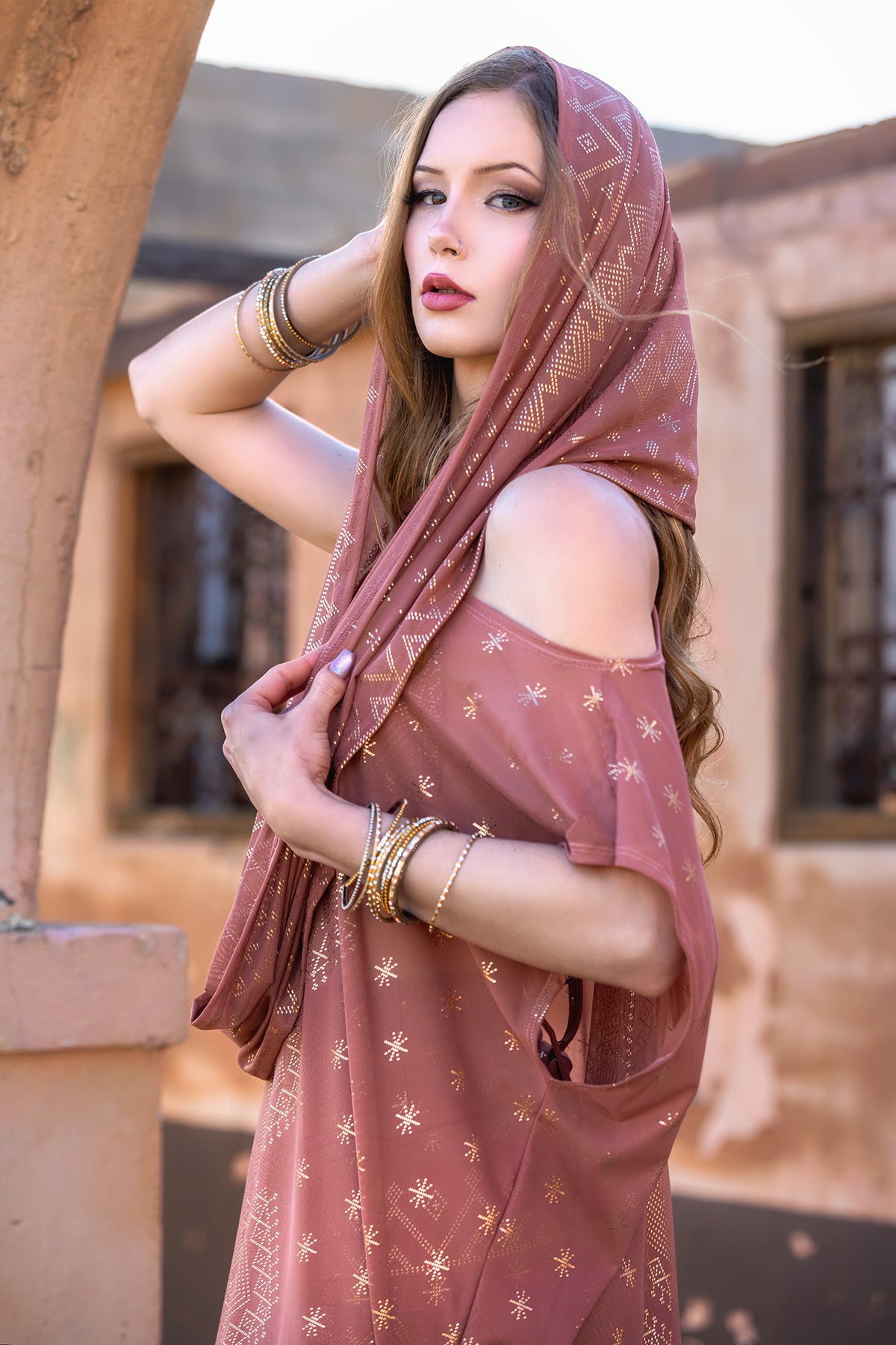 A woman with long brown hair wears Melodia Designs' Dendera Infinity Scarf in Desert Rose, styled with a patterned dress and gold bangles, standing near an arched structure and gazing at the camera with one arm raised.