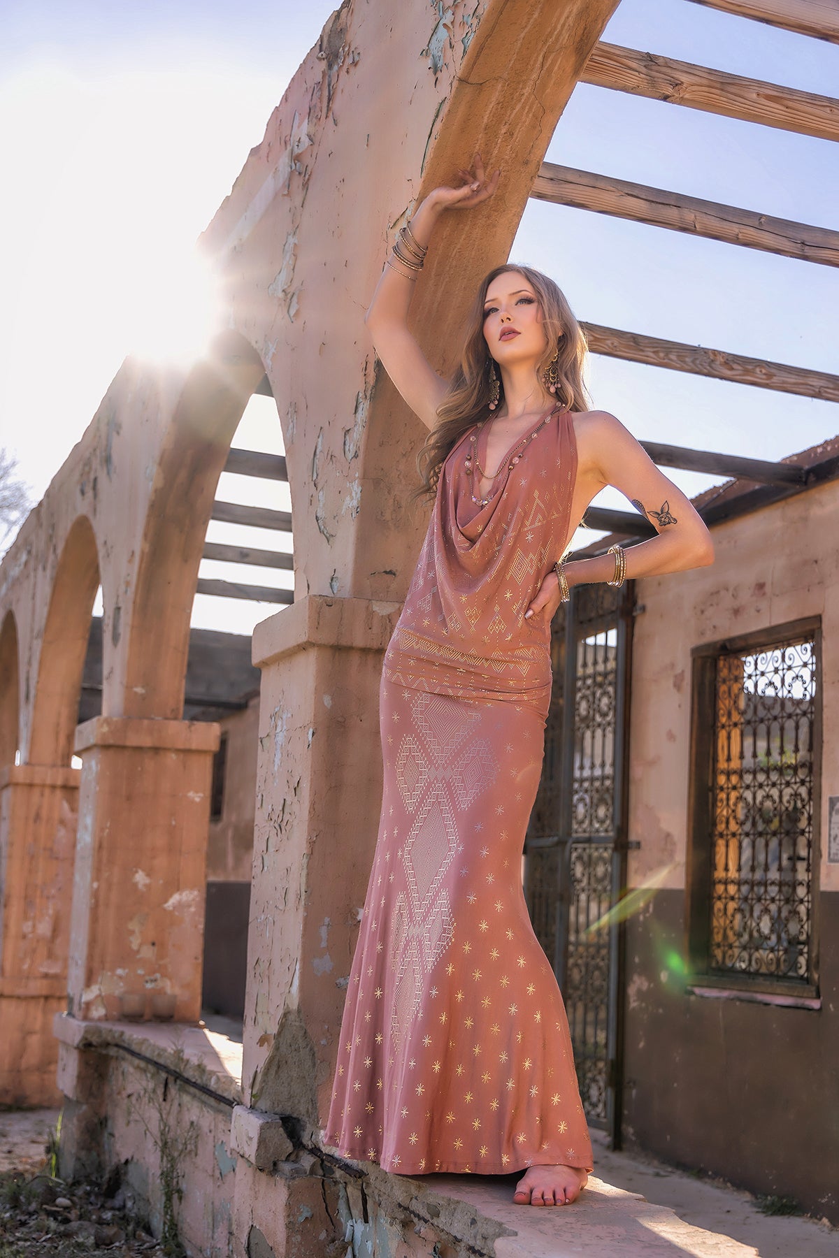 A woman barefoot on a sunlit stone ledge poses in the Dendera Cleopatra Maxi Skirt 2.0 Desert Rose Gold by Melodia Designs, sunlight streaming through open beams above.
