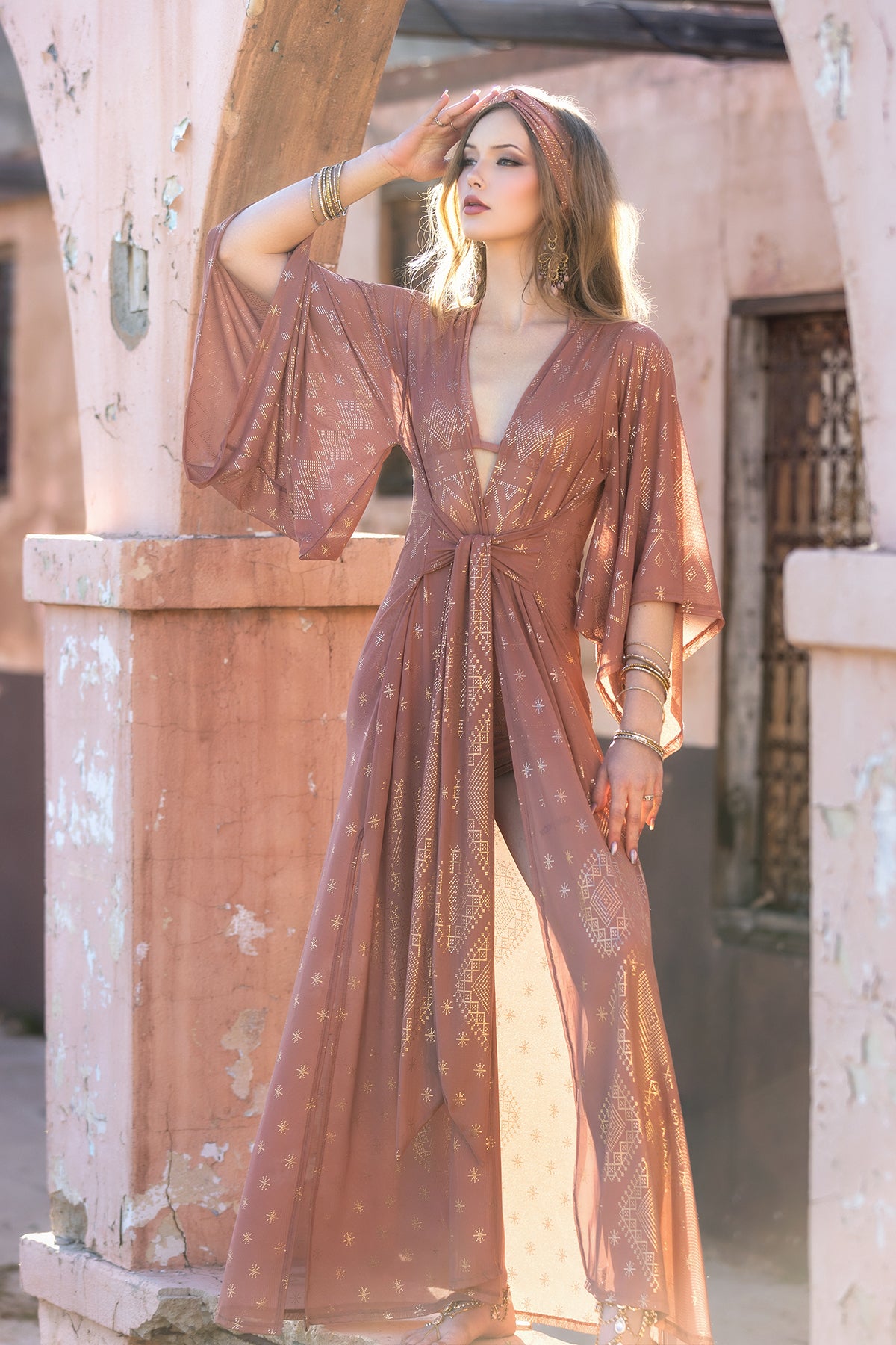 A woman in the Dendera Satori Kimono Desert Rose by Melodia Designs, featuring kimono-inspired sleeves and gold accents, stands under stone arches, gazing into the distance and bathed in warm sunlight.