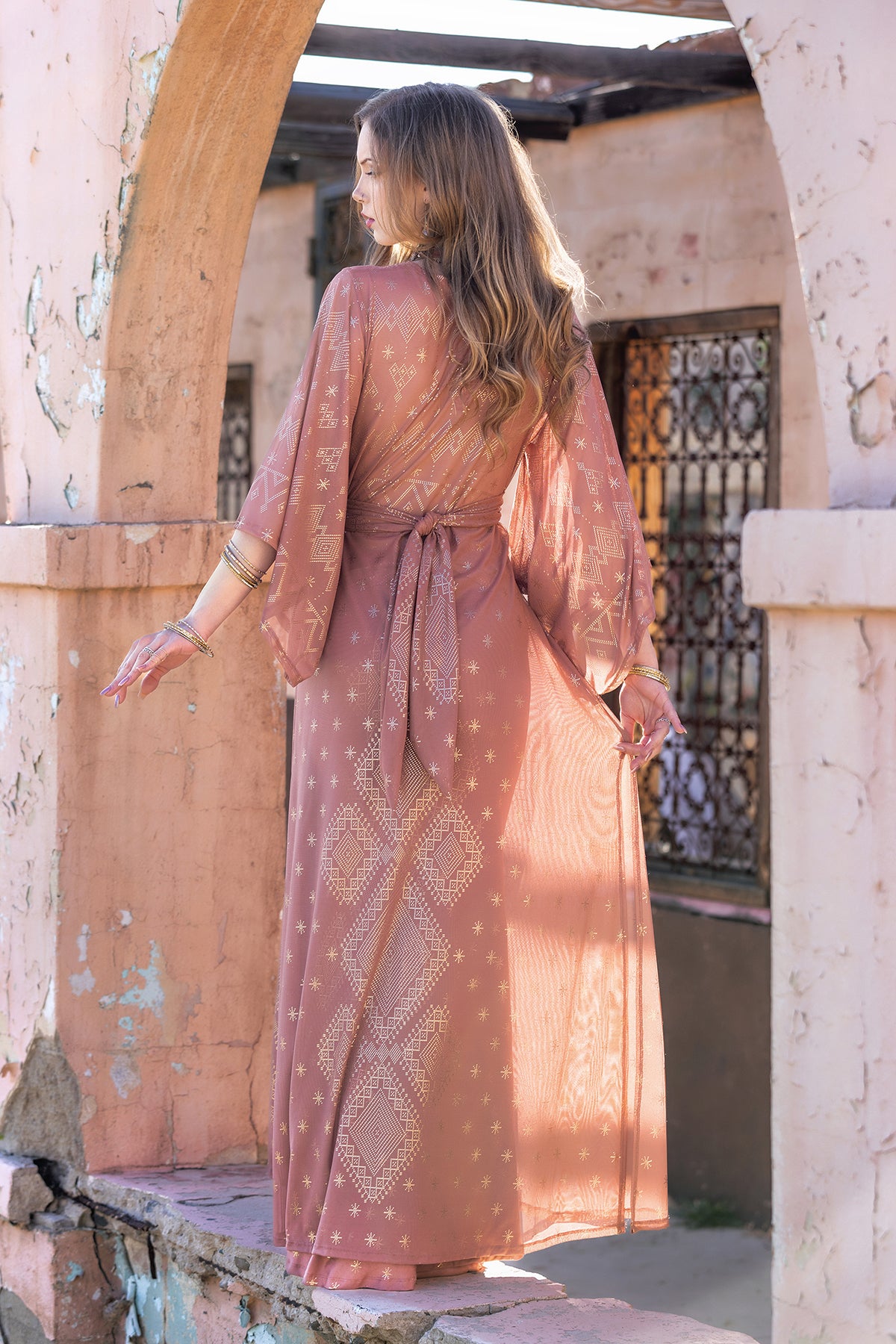 A woman with long wavy hair stands under an archway, facing away. She wears the Dendera Satori Kimono Desert Rose by Melodia Designs—mauve with white geometric patterns, kimono sleeves, and a tie belt—amid rustic pink walls and iron window grilles.