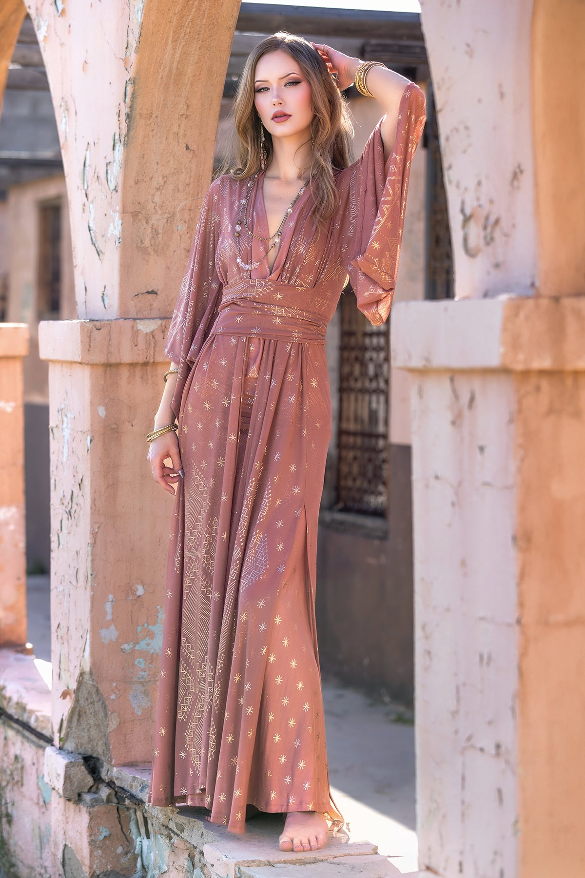 A woman stands barefoot under an arched structure, wearing Melodia Designs' Dendera Satori Kimono Desert Rose with gold star motifs and kimono sleeves. Her loose, wavy hair and gold bracelets complement her confident, serene expression.