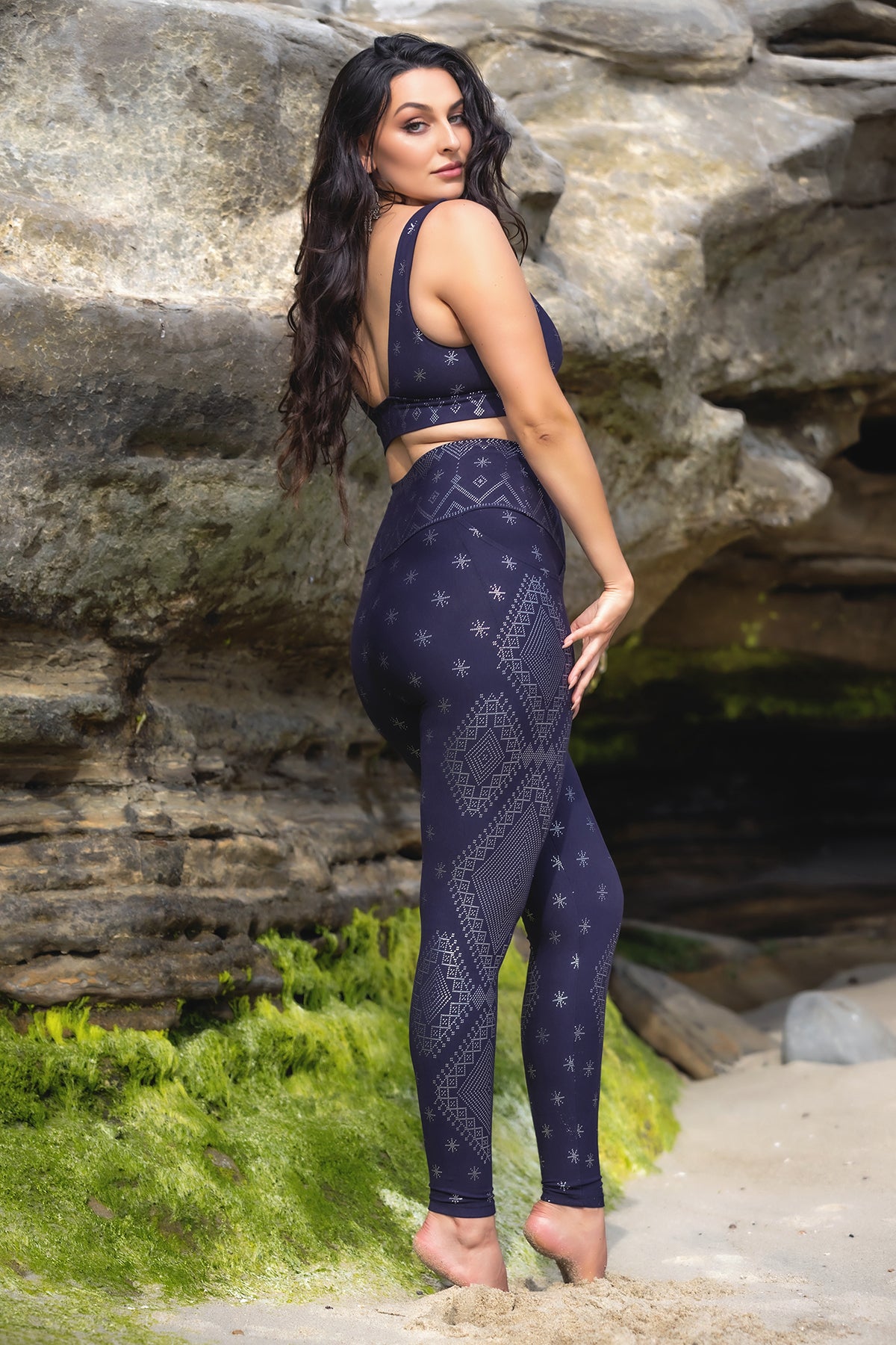 A woman with long dark hair poses barefoot on a sandy beach, wearing Melodia Designs' Dendera Elsa Legging Midnight Silver. The leggings feature a fold-over waistband and geometric star patterns. Rocky cliffs and green moss appear in the background.